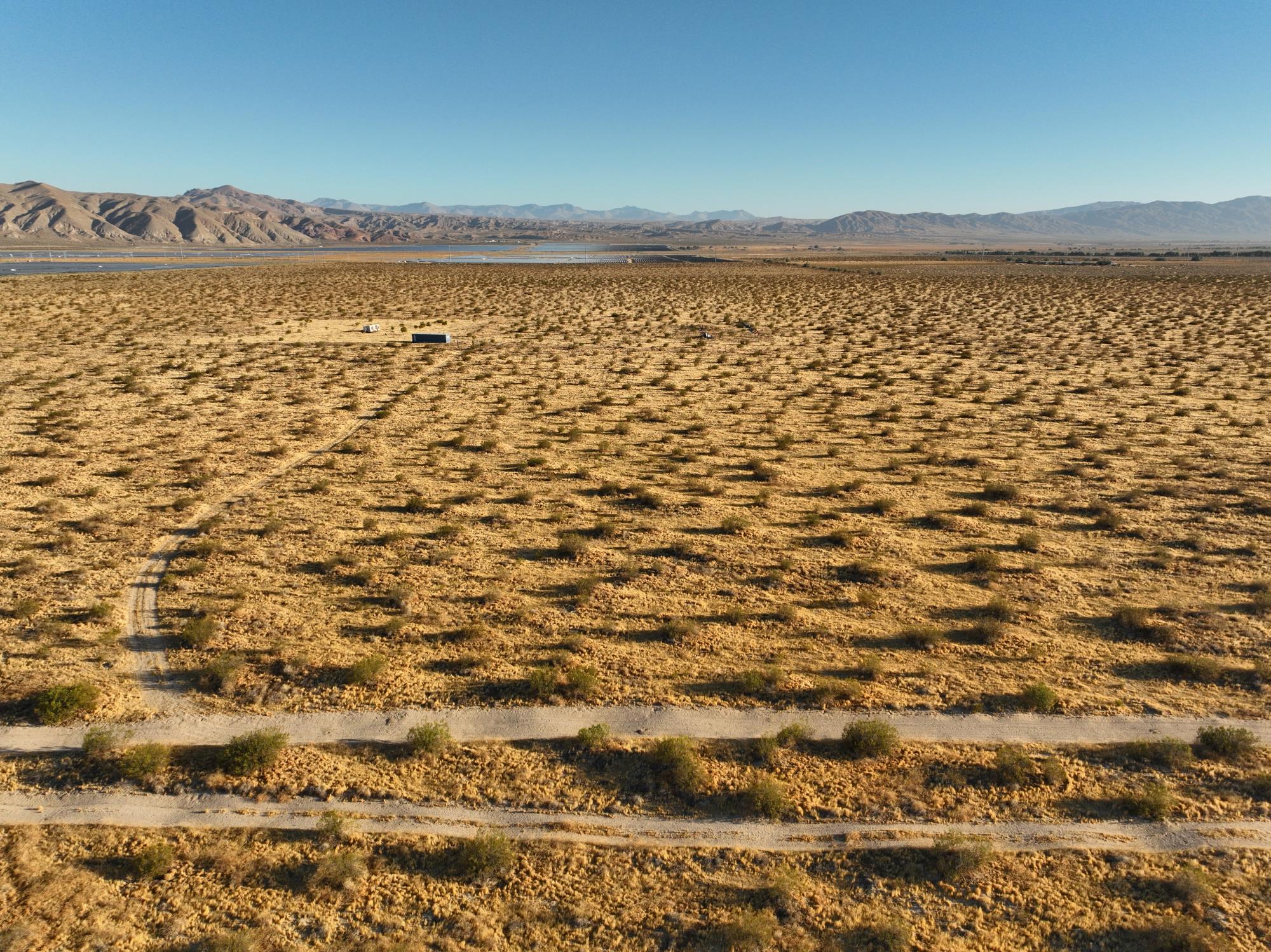 Ariel view of shrubs, and desert grass. A storage container near a dirt path is visible along with metal scrap piles.